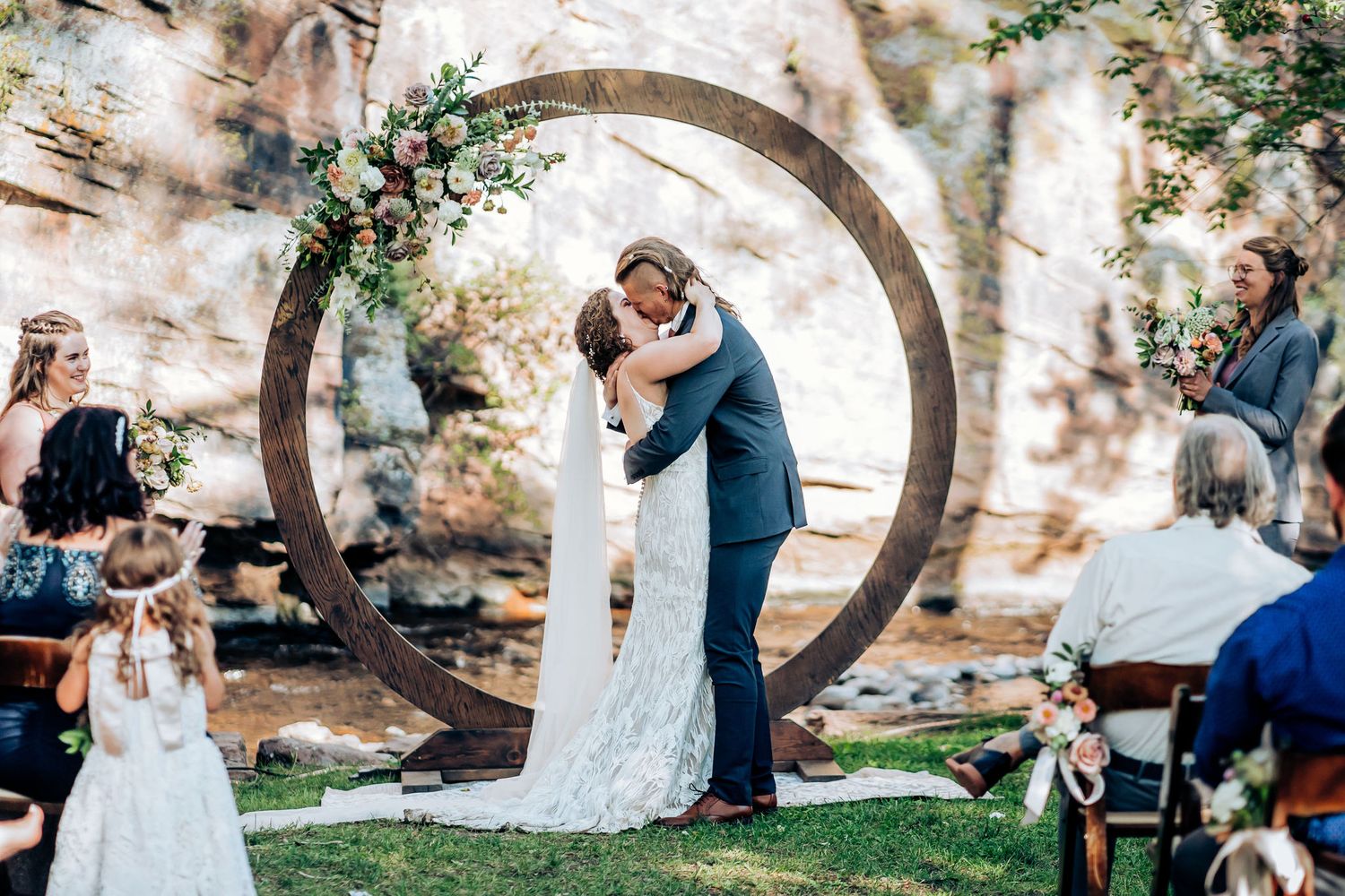 Wedding moment captured under round wooden arch adorned with greenery against stone wall backdrop.