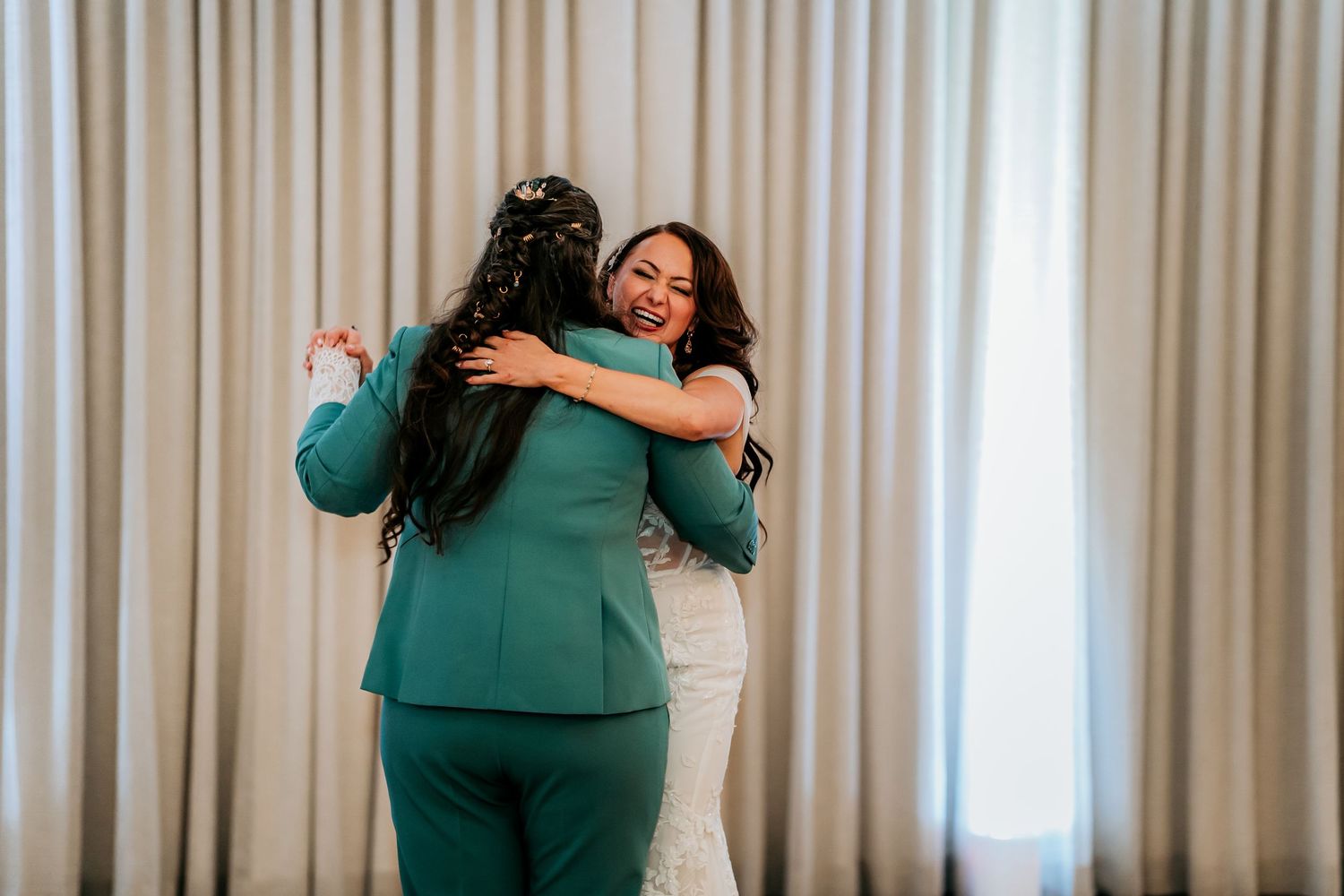 Two brides share a joyful embrace on the dance floor in a white lace gown and teal suit.