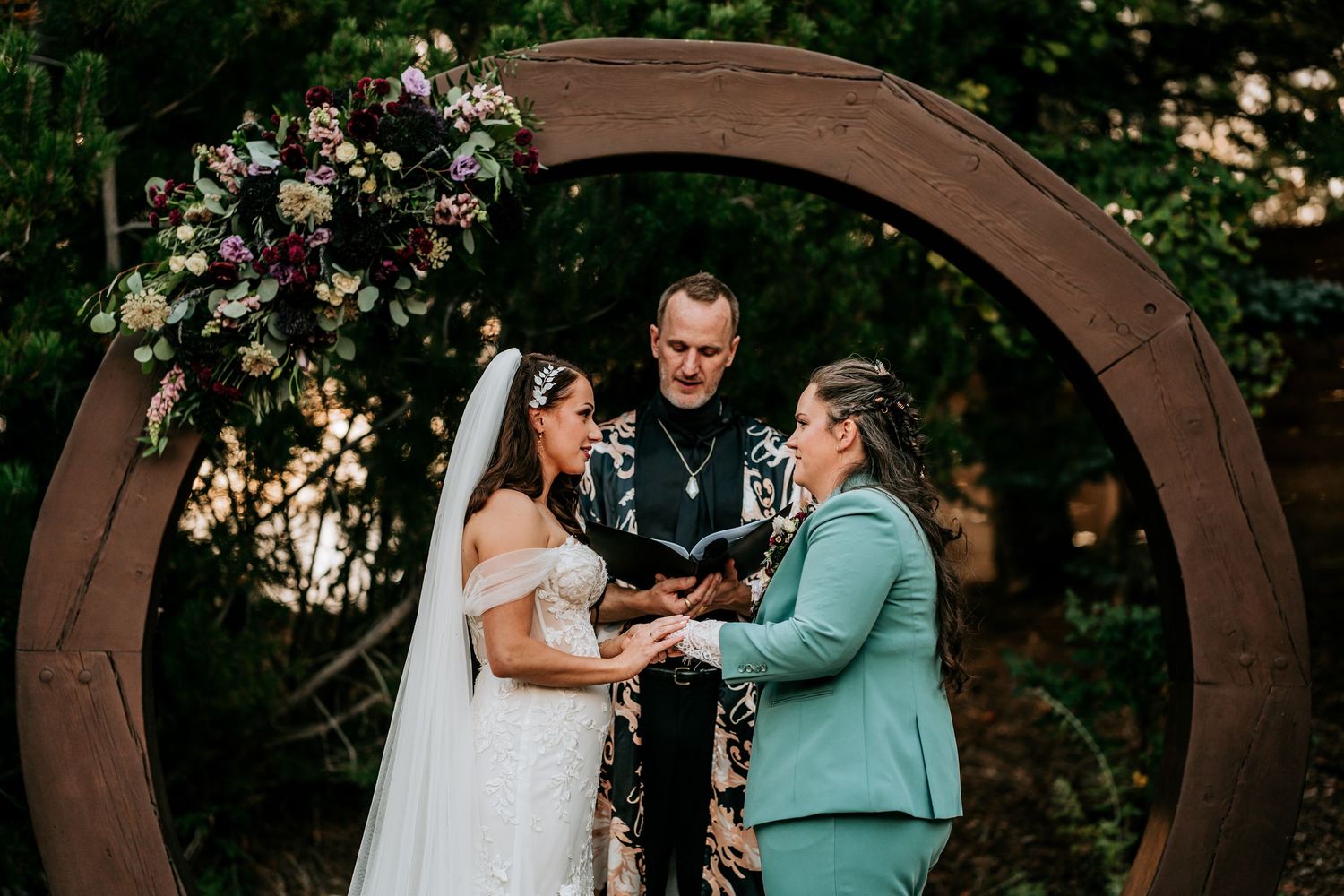 Two brides stand with officiant under a circular arch with lush floral arrangements at outdoor wedding.
