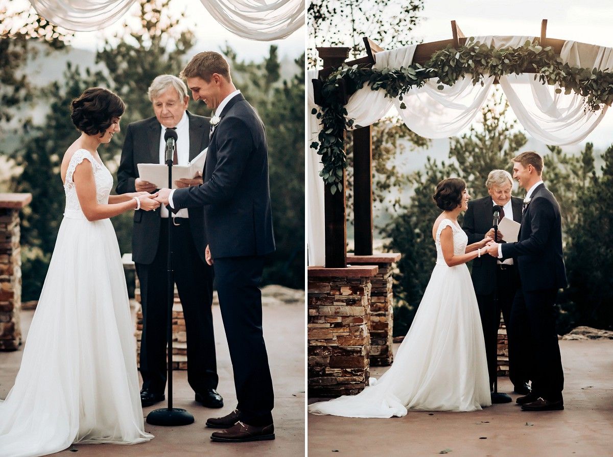 Wedding ceremony taking place under stone pillars and white draping with evergreen trees in background.