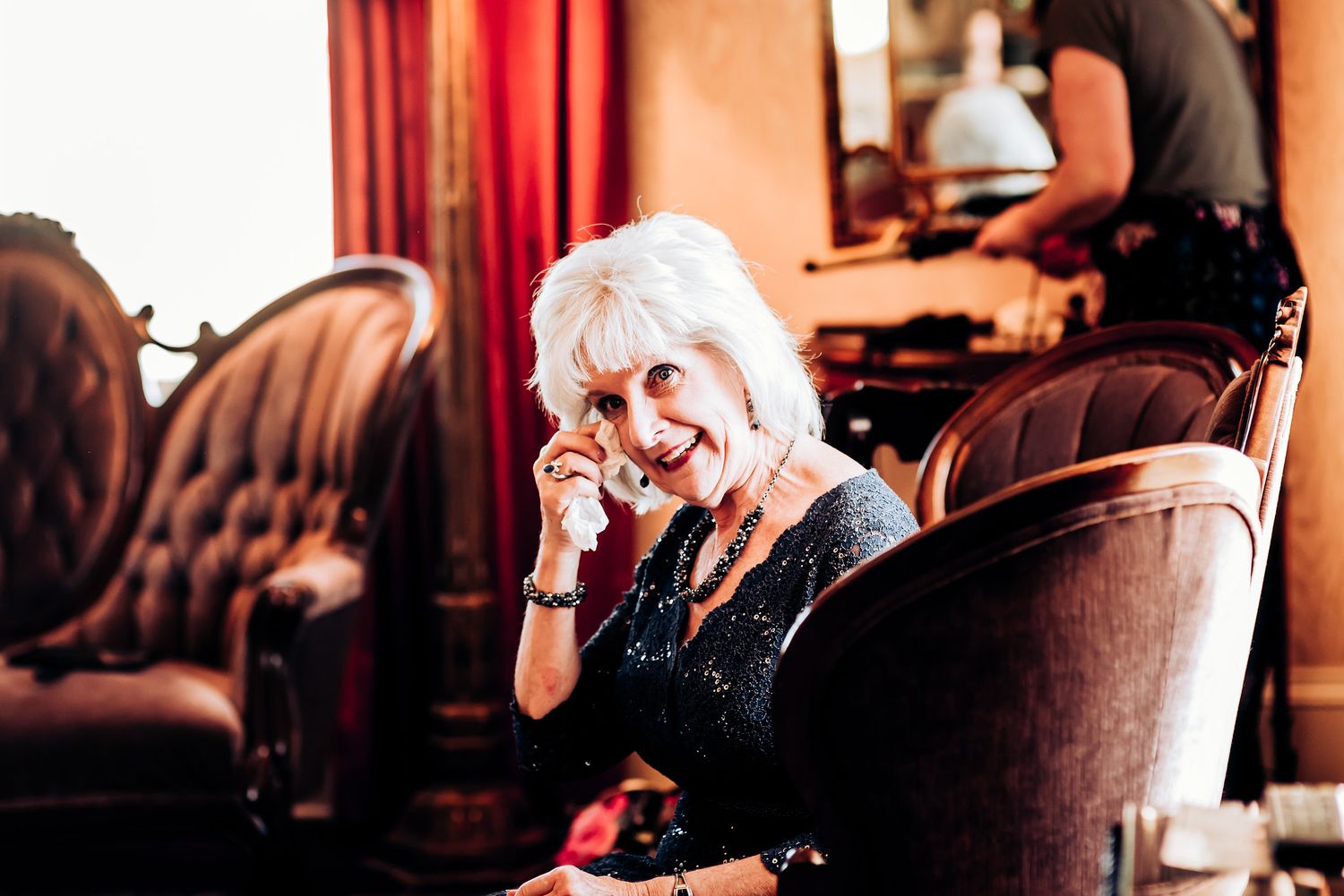 An elderly woman in a black outfit laughs while sitting in an elegant armchair at an indoor venue.