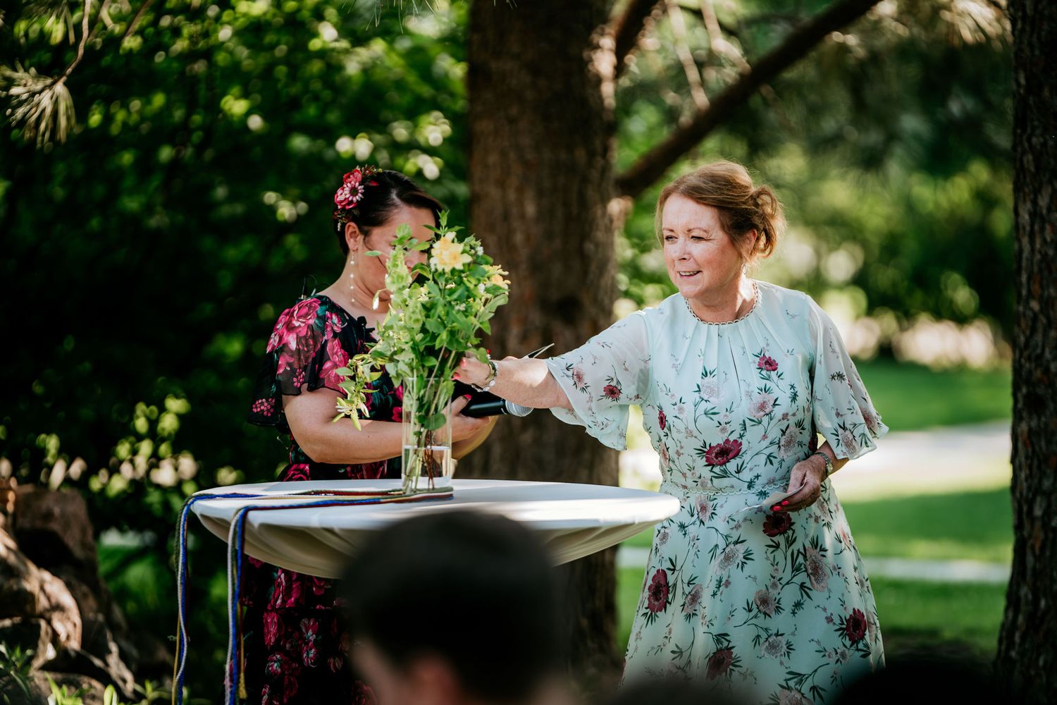 Two people in floral dresses arrange flowers on a white table outdoors.