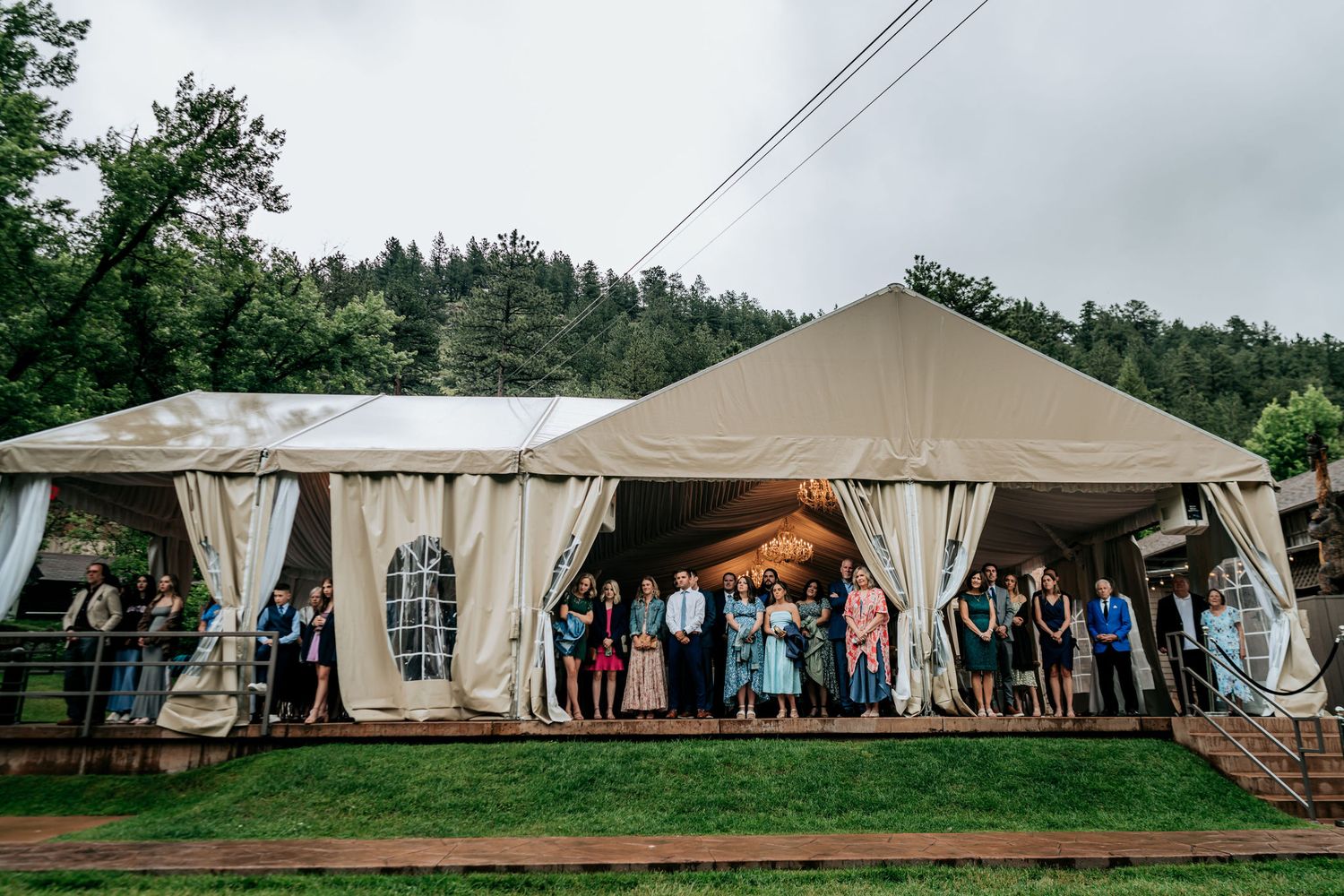 Large outdoor wedding reception tent with white drapes and guests mingling under tan canvas in mountain setting.