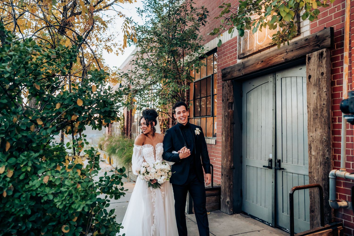 Newlyweds walk together along brick wall with greenery, bride holding white floral bouquet.