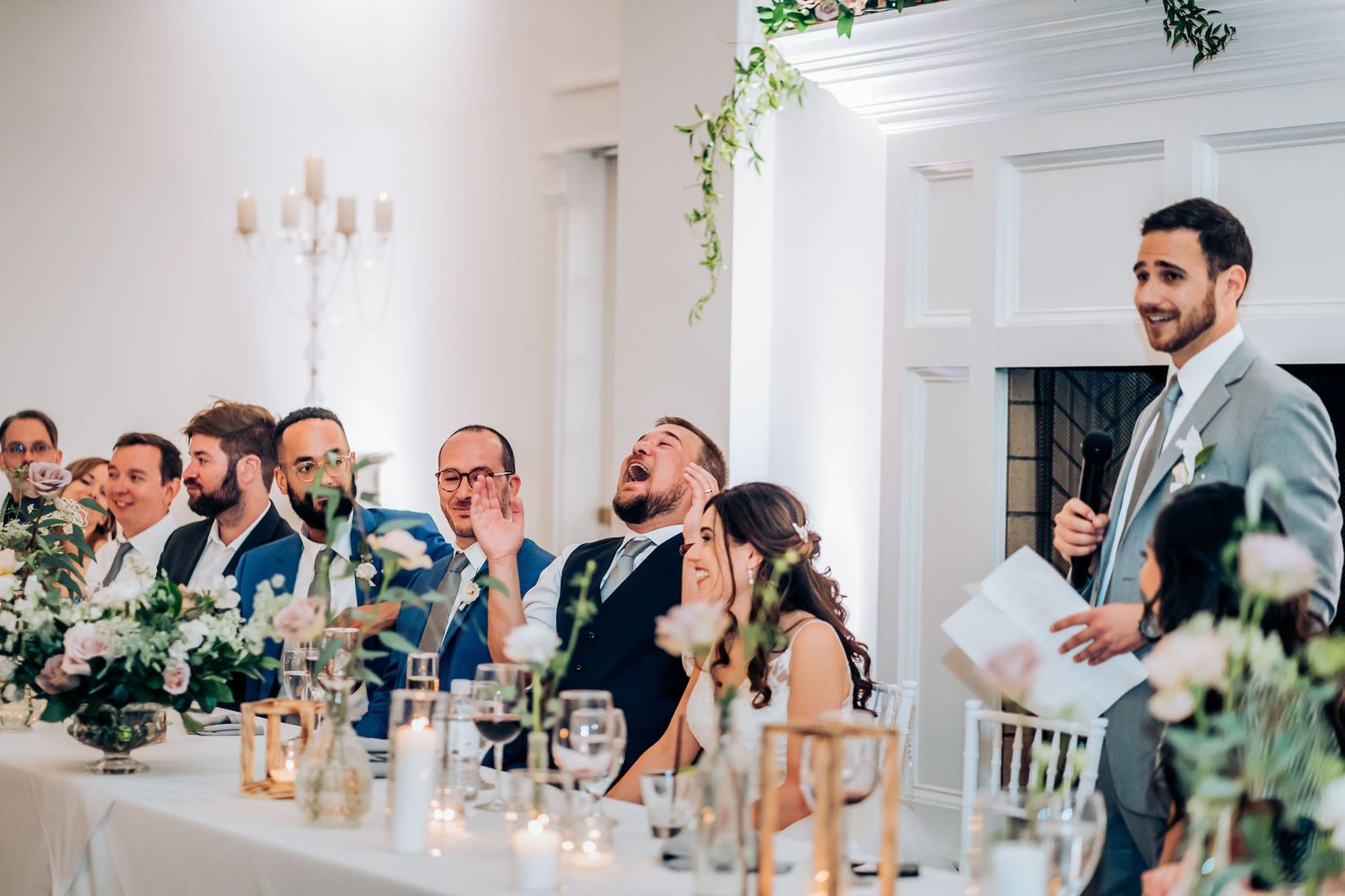 Wedding party members laugh and react during reception speeches at decorated head table.