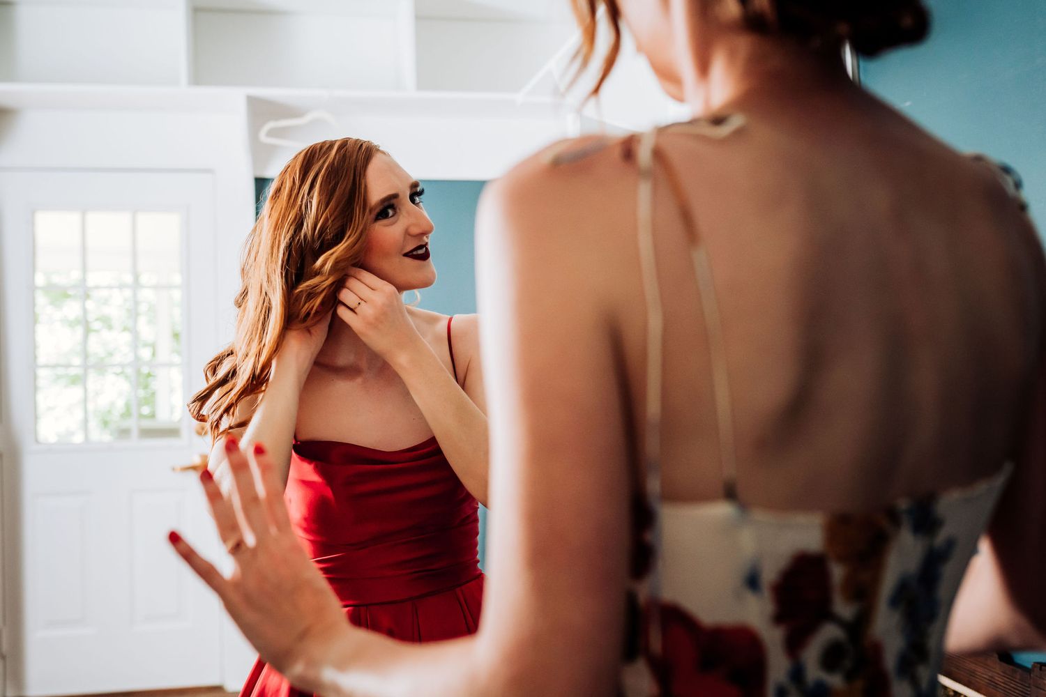 Woman in red dress looks at reflection while getting ready in bright interior space.