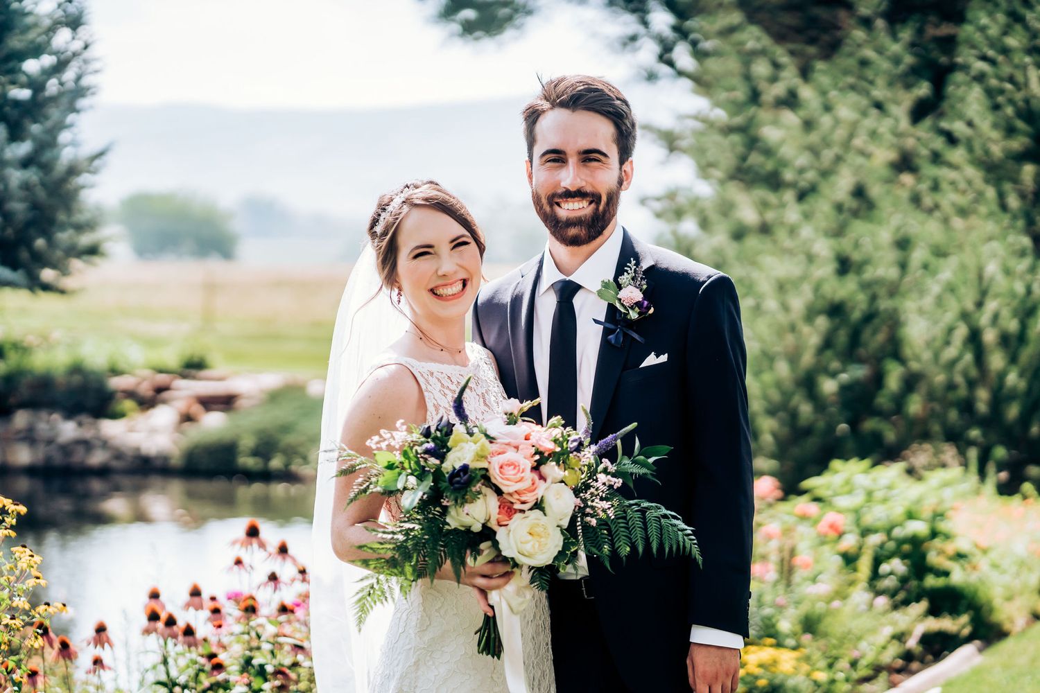 Newlyweds pose together in garden with pink flowers and water feature in background.