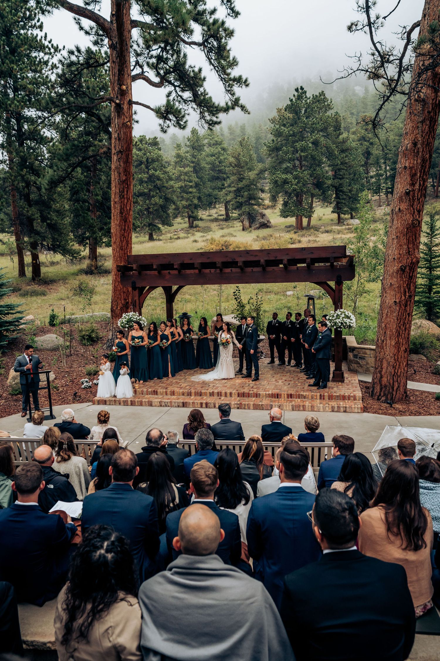 Wedding guests watch a ceremony from wooden chairs as fog rolls through the pine forest backdrop.