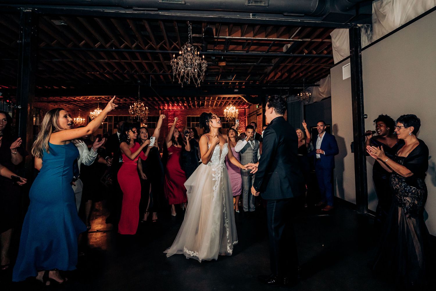 Wedding guests and bride dance beneath chandeliers at a dimly lit reception venue.
