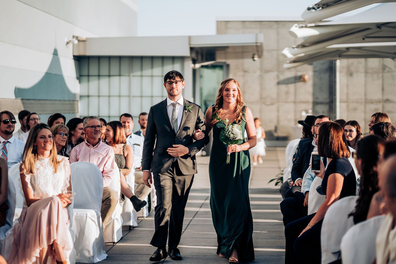 Wedding party members in formal attire process down an aisle between seated guests at an outdoor venue.
