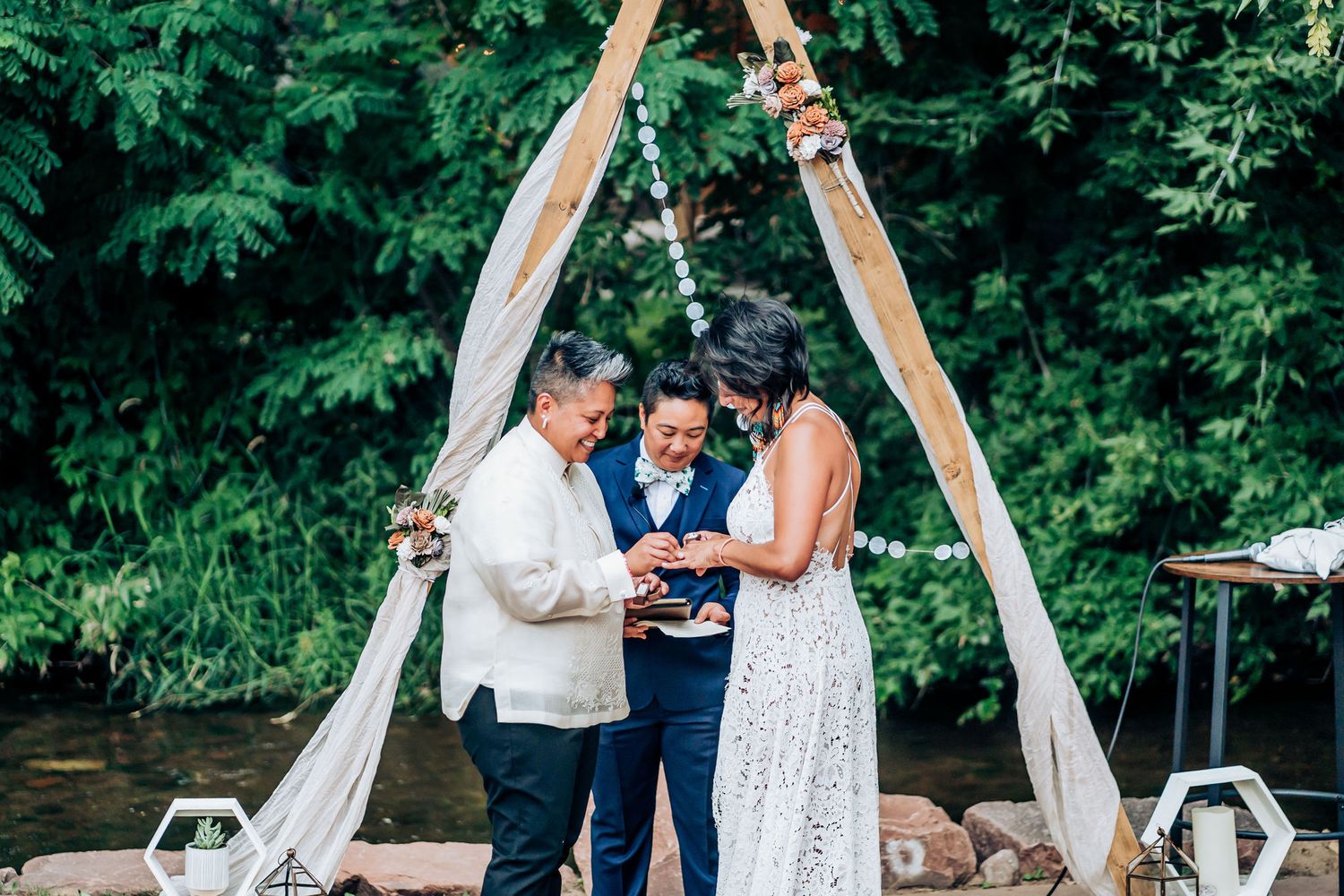 An outdoor wedding ceremony takes place under a triangular wooden arch with white fabric and floral accents.
