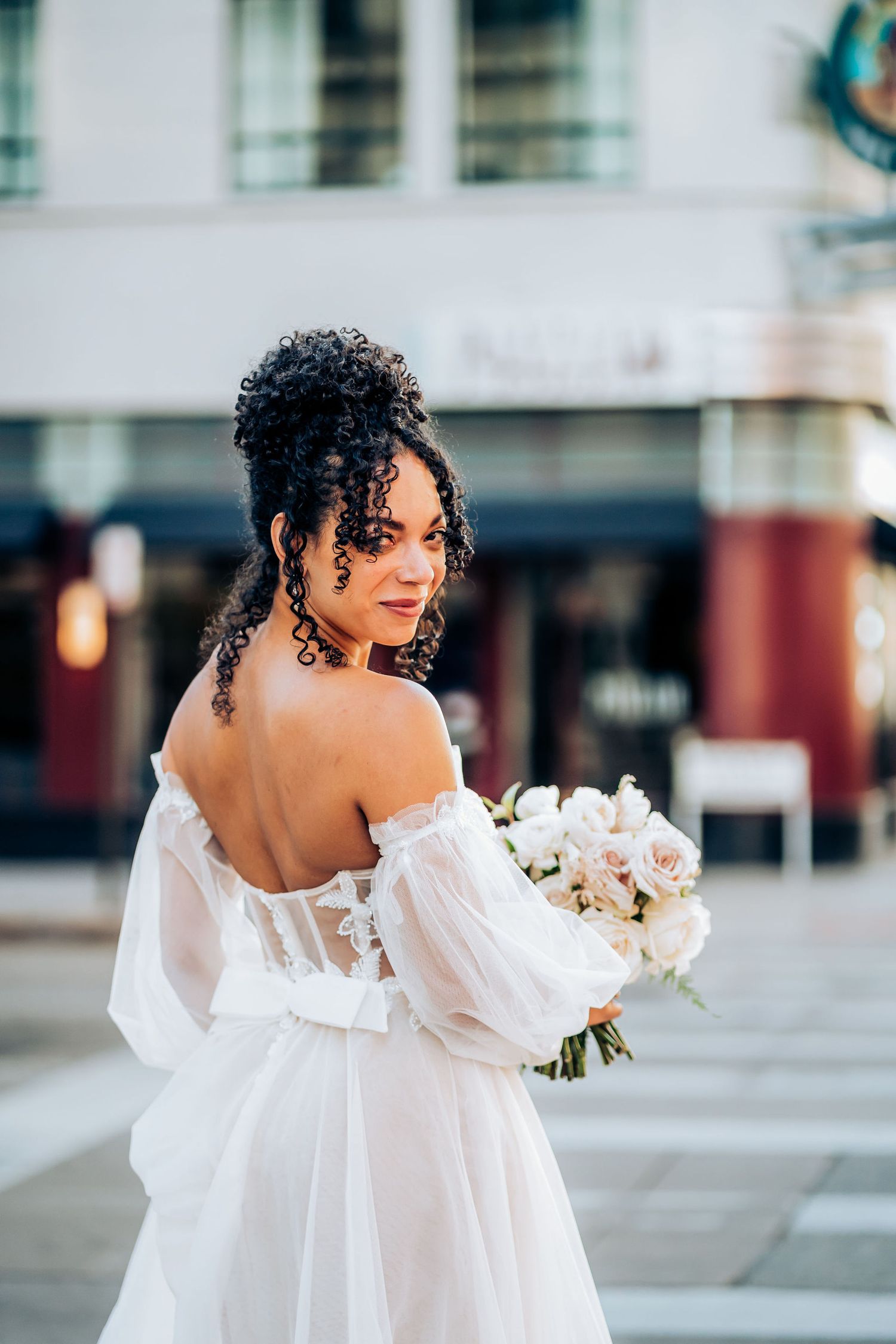 Bride in off-shoulder wedding gown holds white bouquet while looking over shoulder.