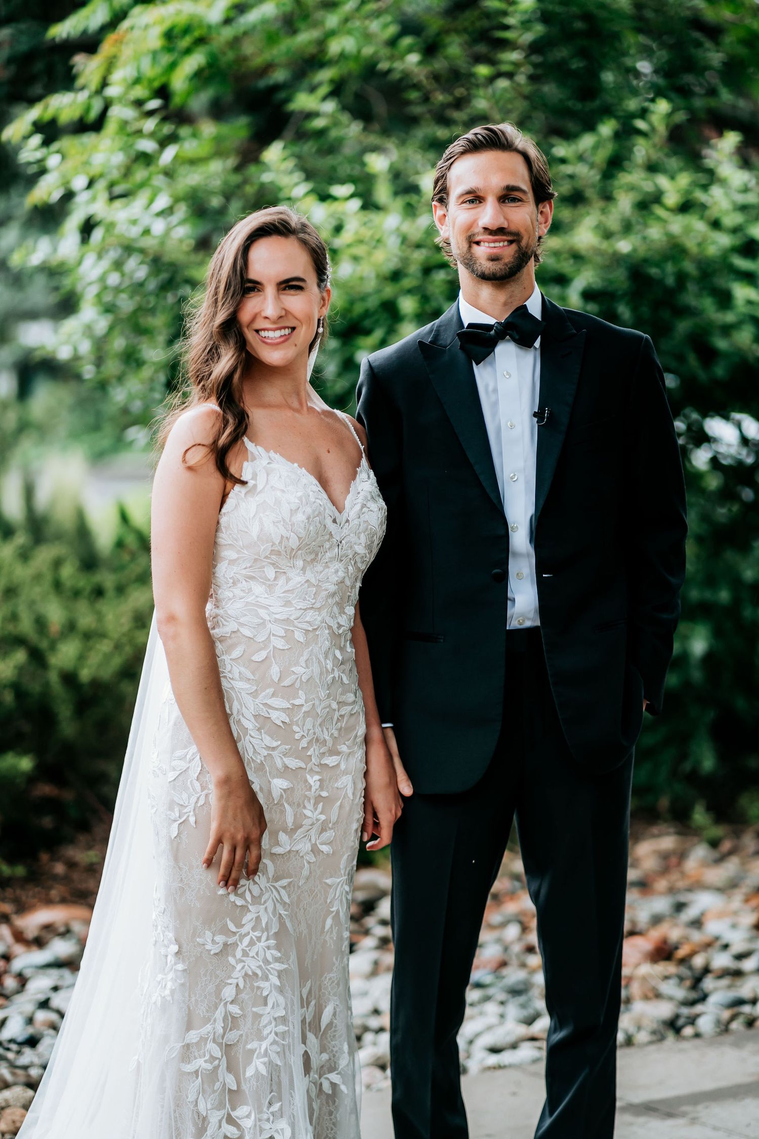 A newlywed couple poses outdoors in formal wedding attire against a leafy green backdrop.