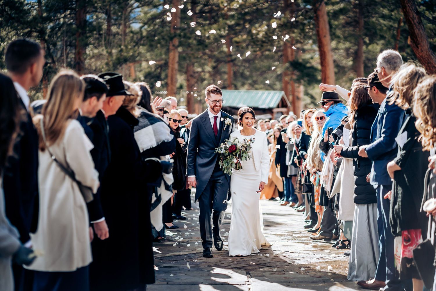 Wedding exit as couple walks down tree-lined path with guests taking photos.