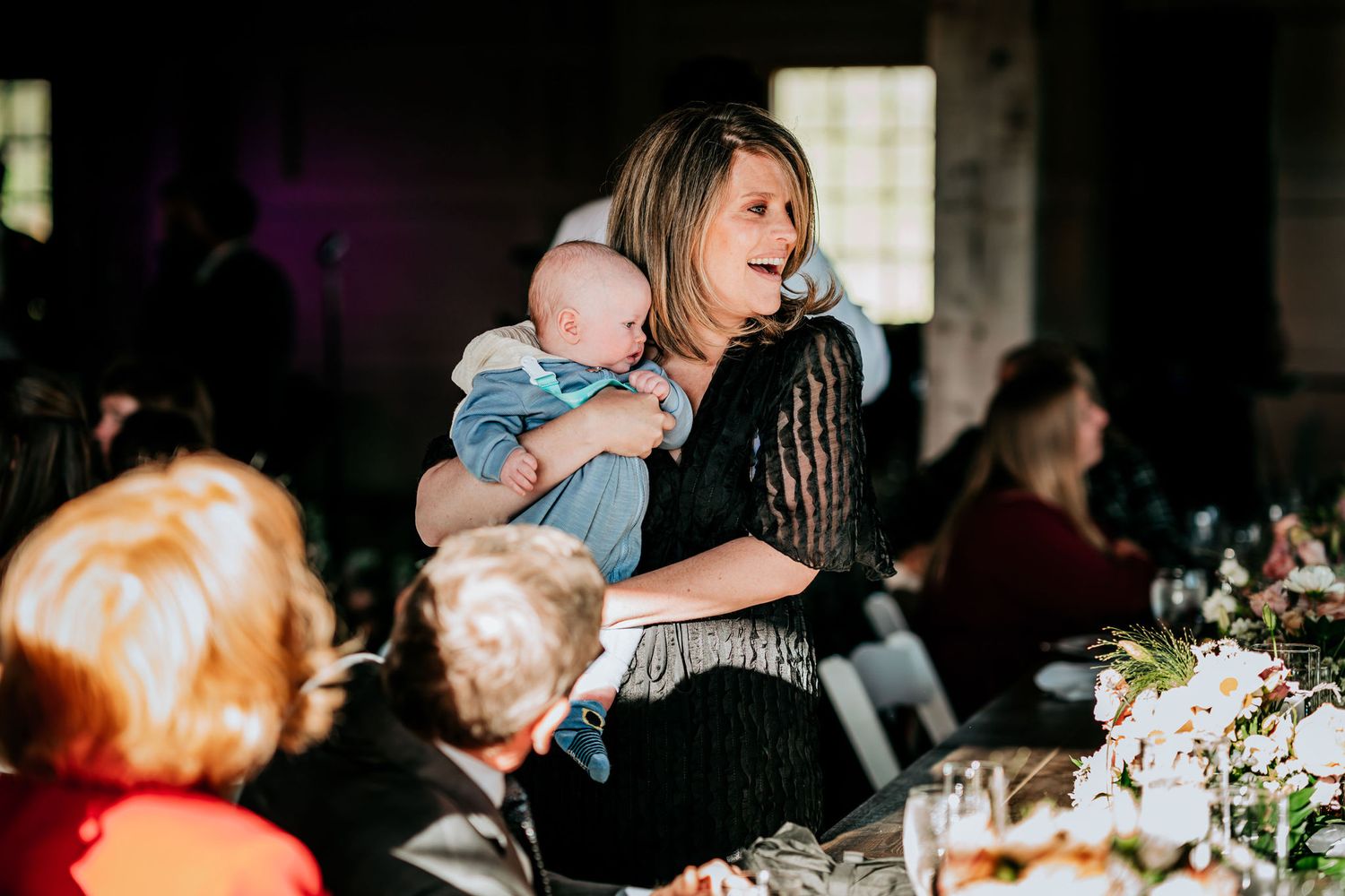 A guest holds a baby during a wedding reception dinner in a rustic venue.