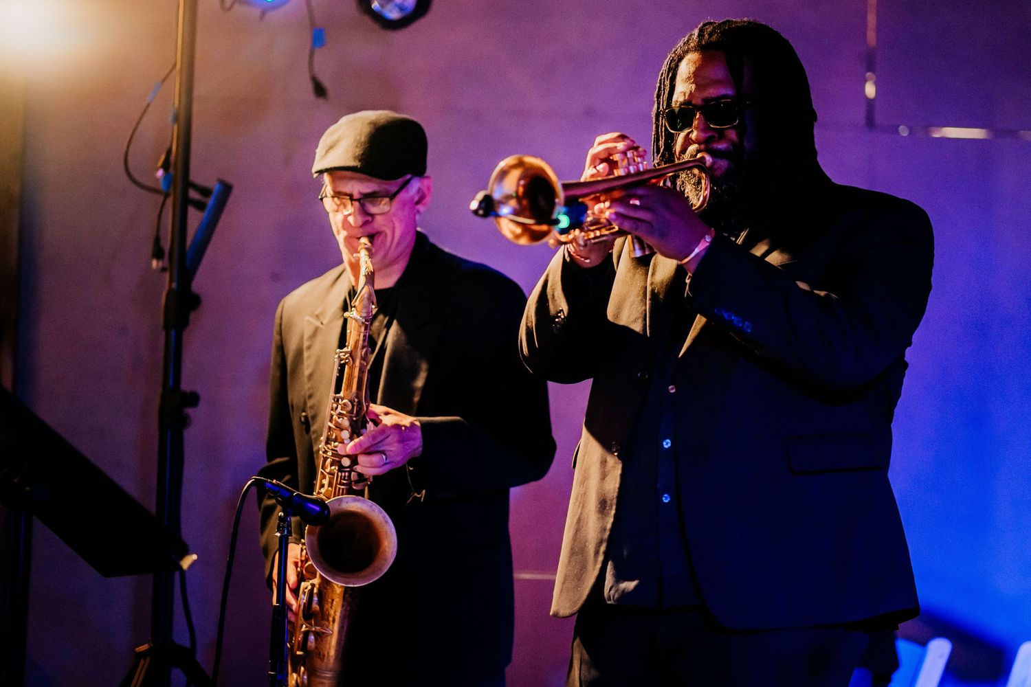 Musicians perform with saxophone and trumpet against blue and purple stage lighting.