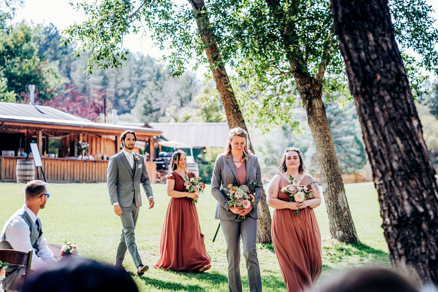 Wedding party members in gray suits and burgundy dresses standing under trees during outdoor ceremony.