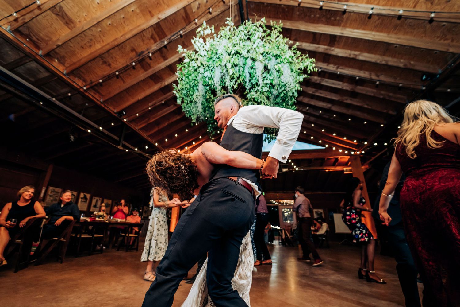 Dancers perform energetic dance moves during a reception under hanging string lights and greenery.
