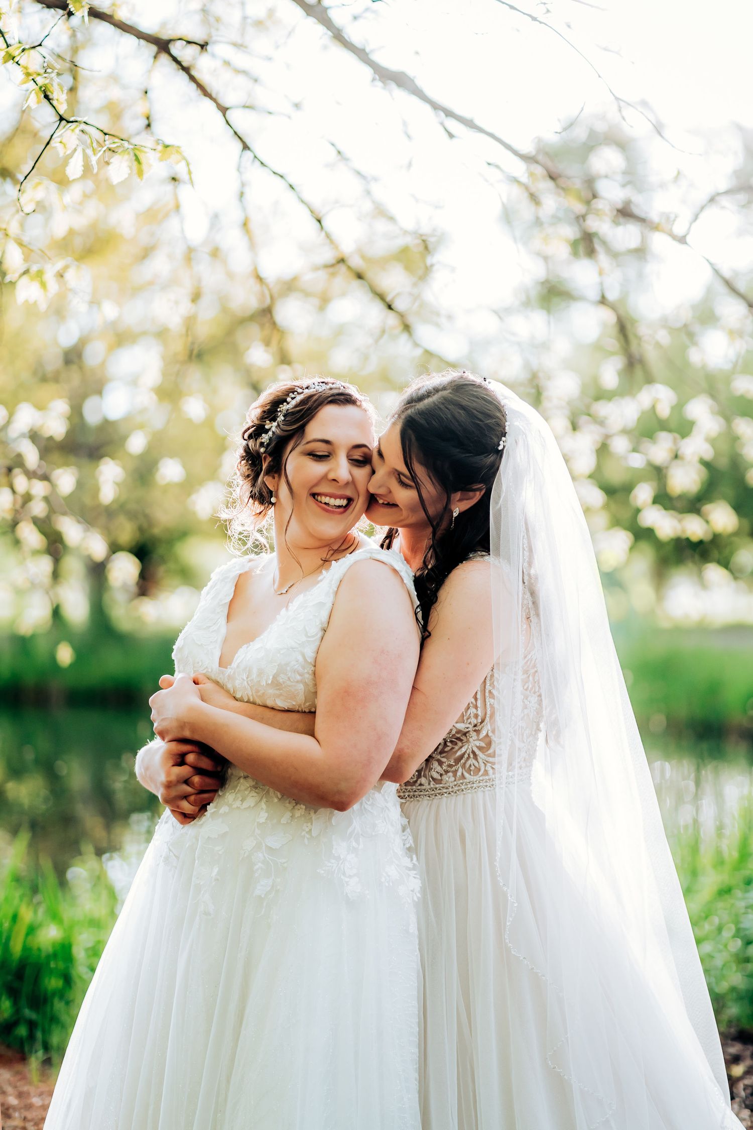 Wedding couple sharing a tender embrace in white gowns surrounded by spring greenery.