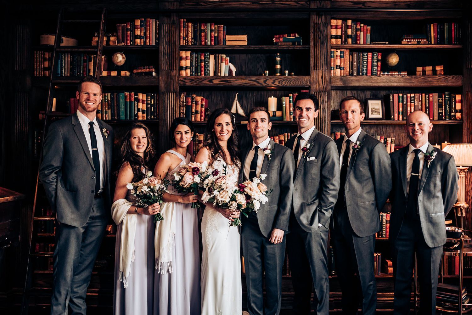 Wedding party in formal attire poses against dark wood library shelves filled with vintage books.