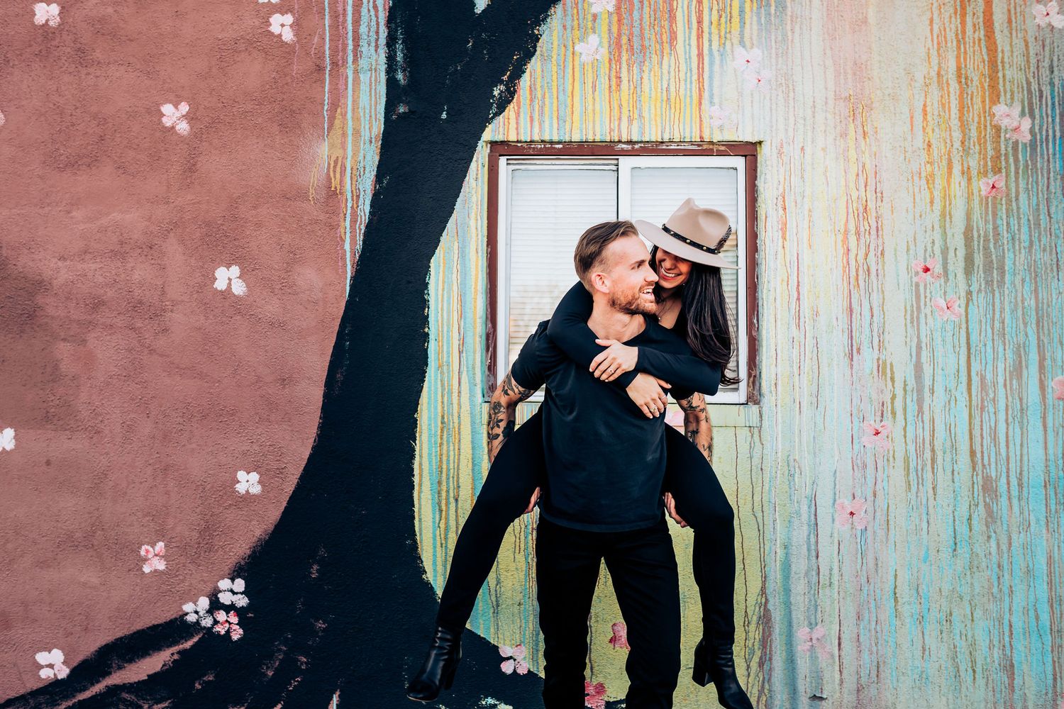 Couple plays and laughs in front of weathered wall with window frame.