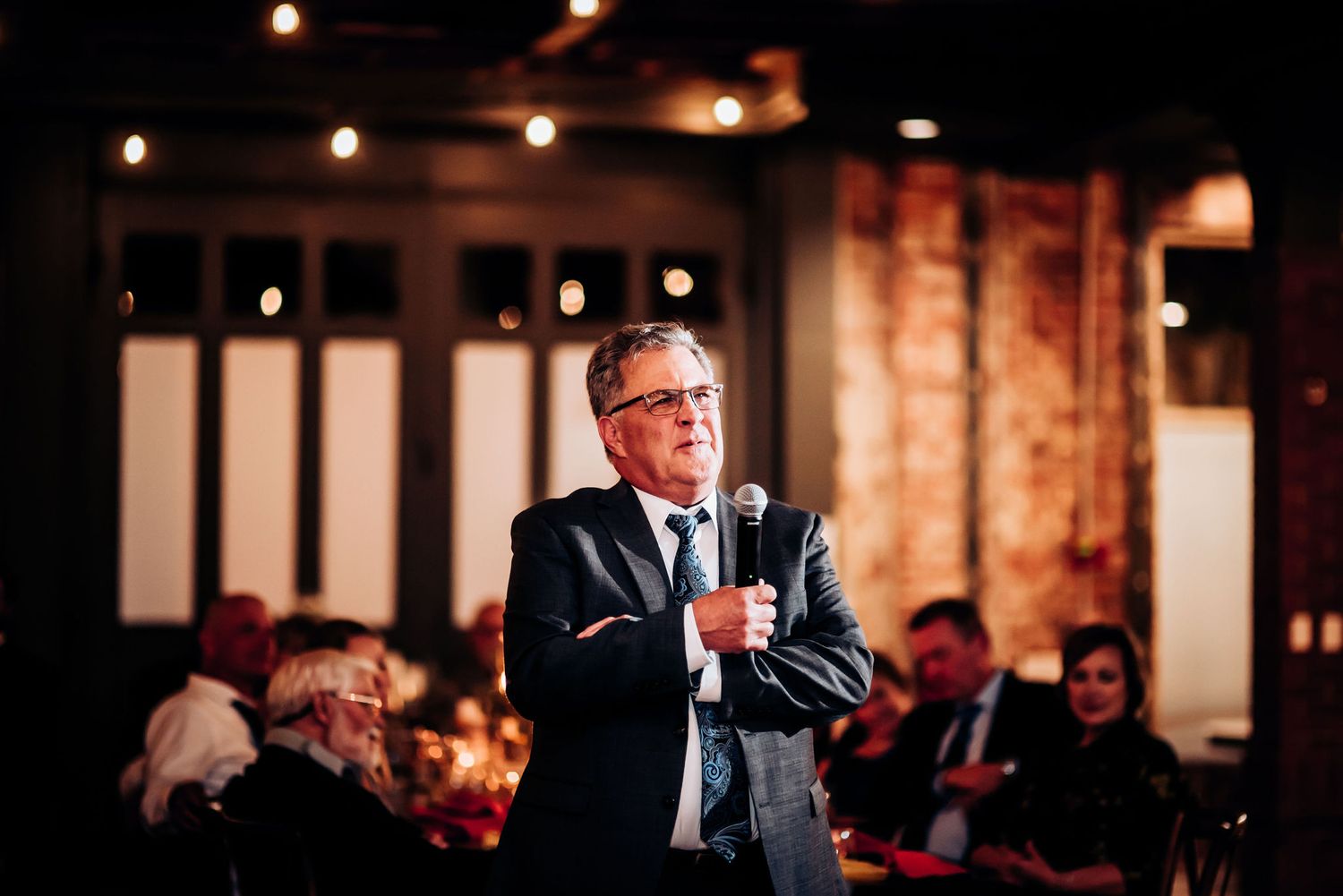 A speaker presenting to an audience in a warmly lit venue with exposed brick walls.