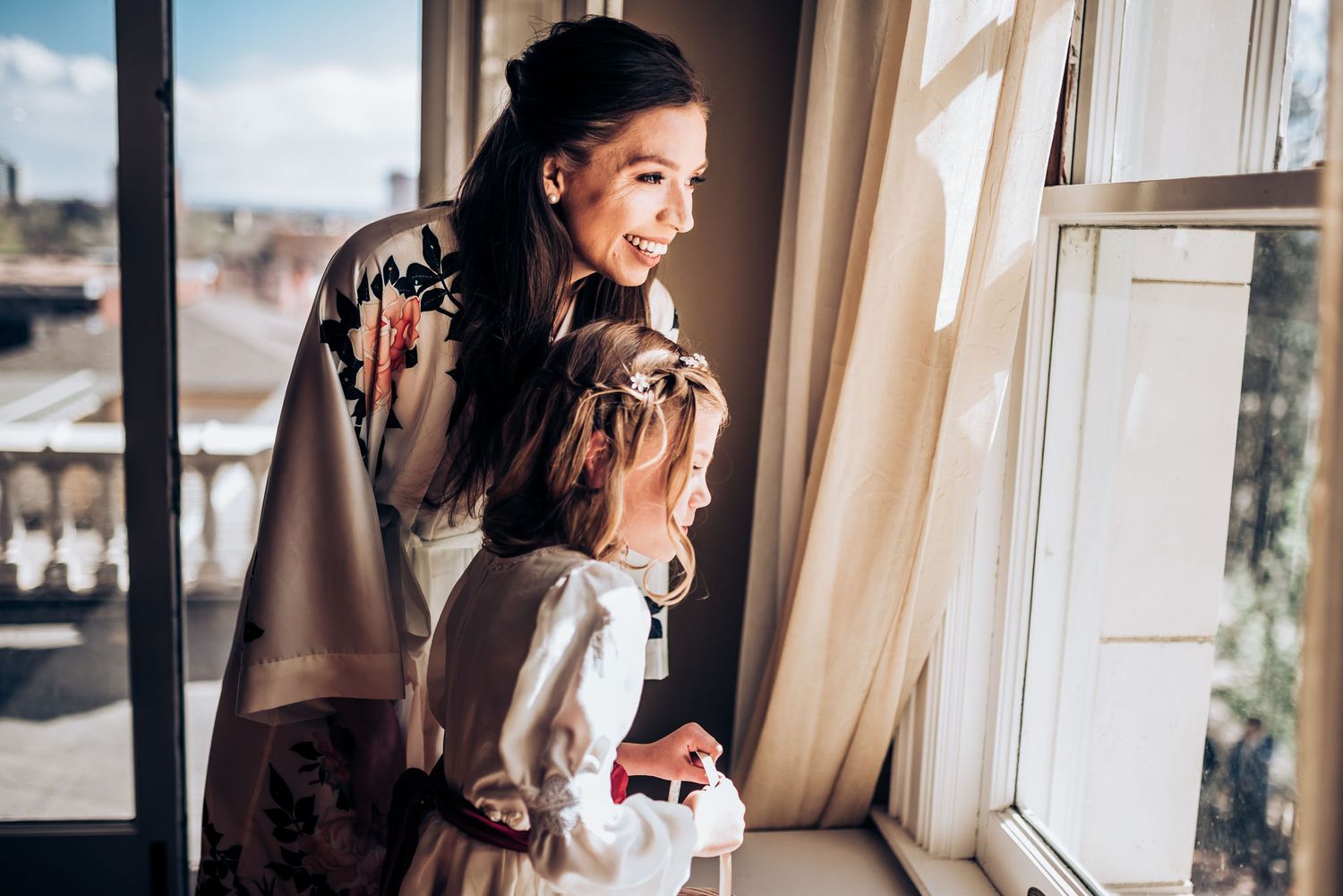 Two figures sharing a joyful moment by a bright window during wedding preparations.