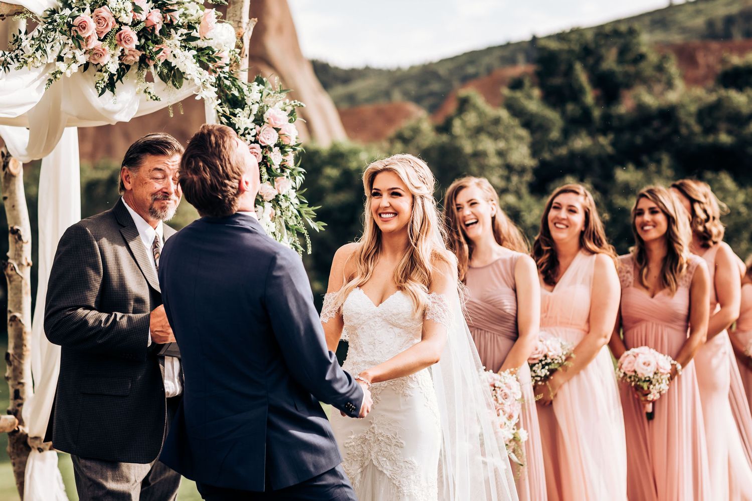 Wedding ceremony with bridal party in pink dresses standing beside decorated wooden arch.