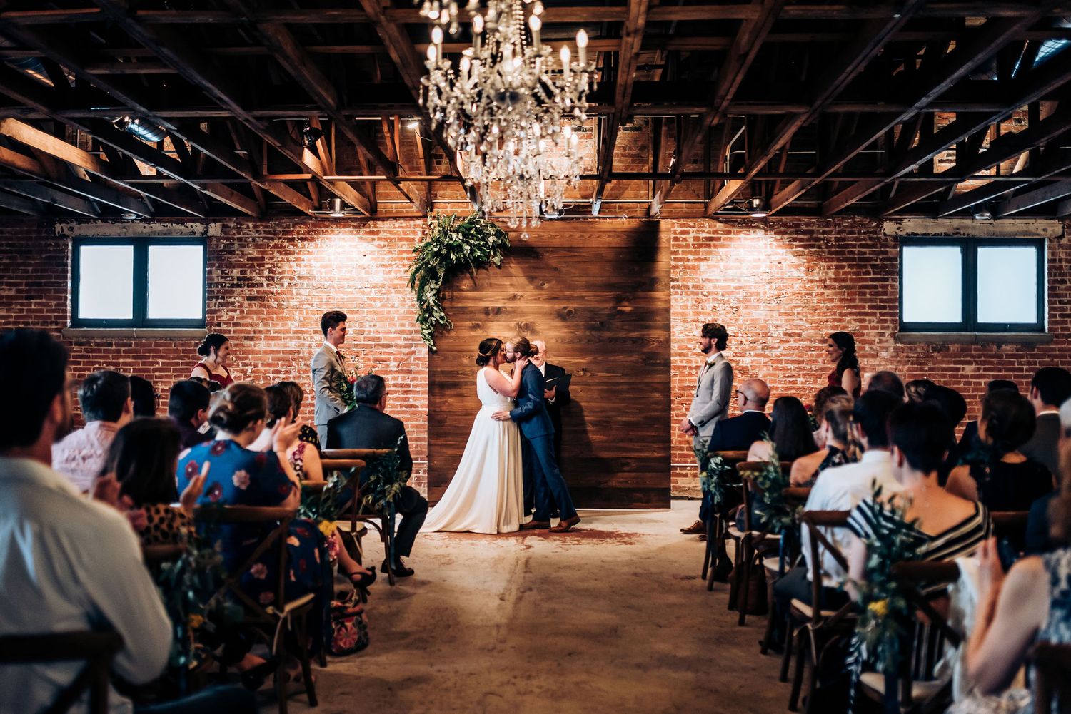 Wedding ceremony in rustic venue with elegant chandelier and wooden accent wall as guests look on.