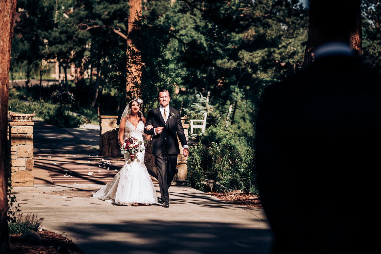 A couple in formal wedding attire walks along a stone bridge pathway surrounded by pine trees.