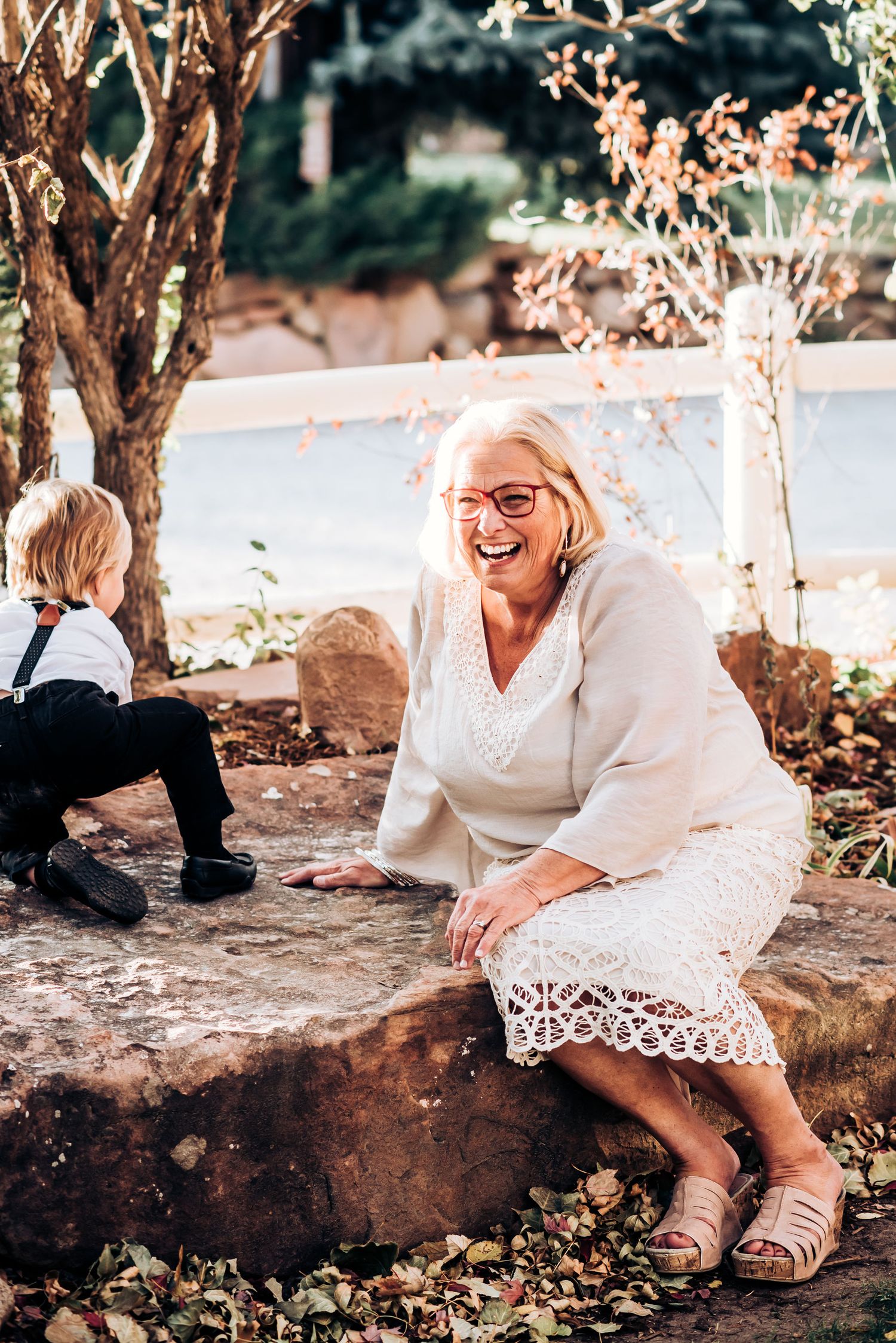 A woman in white dress sitting on stone wall in garden setting with flowering trees.