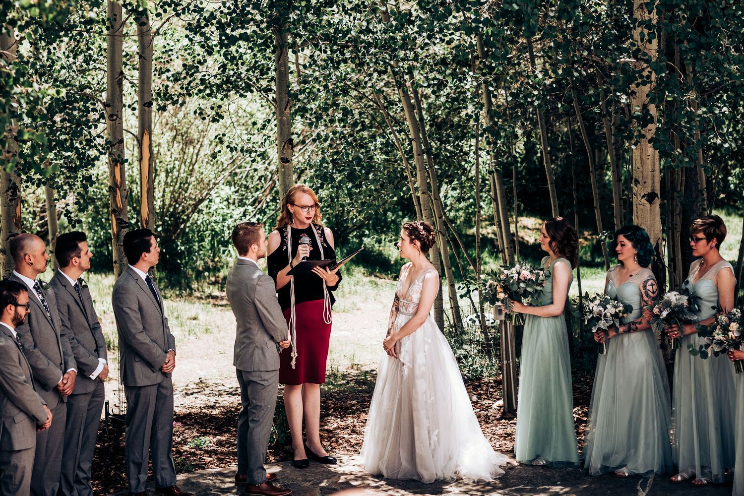 A wedding ceremony in a wooded setting with the wedding party lined up facing the officiant in red.