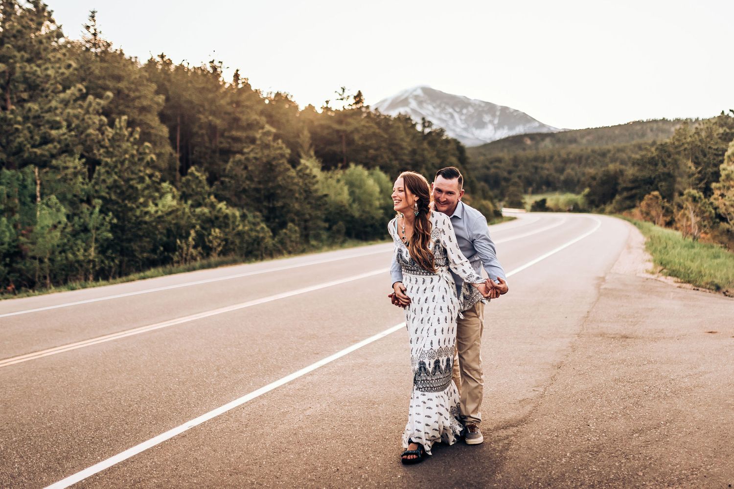 A couple embraces on a scenic mountain road during sunset with tall trees in the background.
