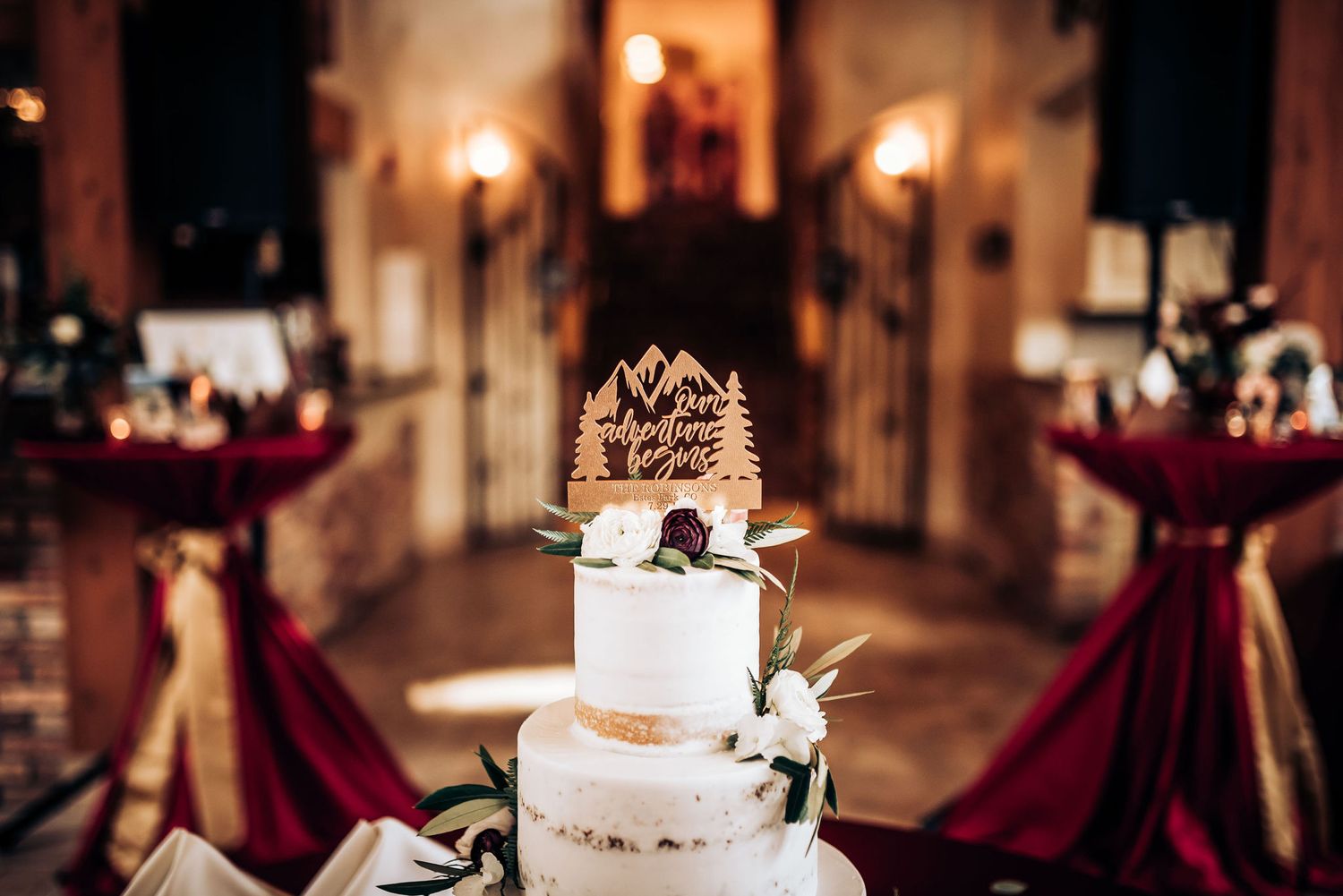 Elegant white wedding cake with burgundy accents displayed in a romantic venue with red table settings.