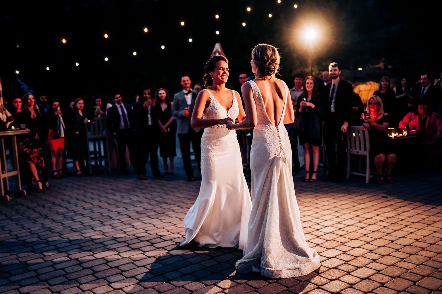 Two brides in elegant white wedding gowns share their first dance on a brick patio under evening lights.
