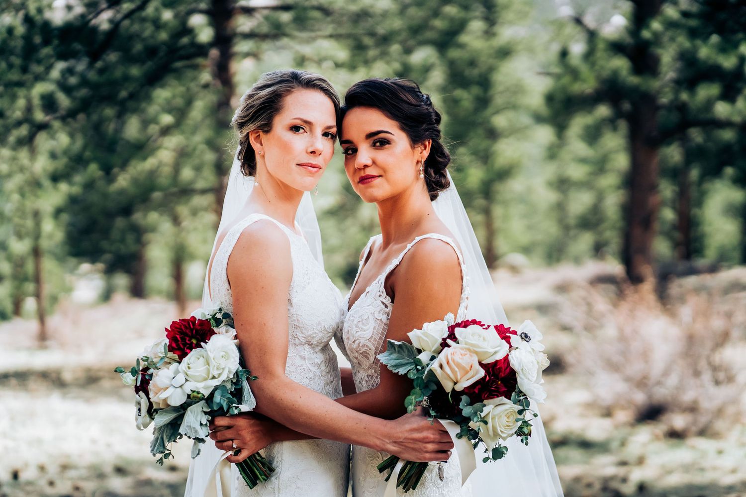 Two brides hold white and pink floral bouquets while posing together outdoors in a forest setting.