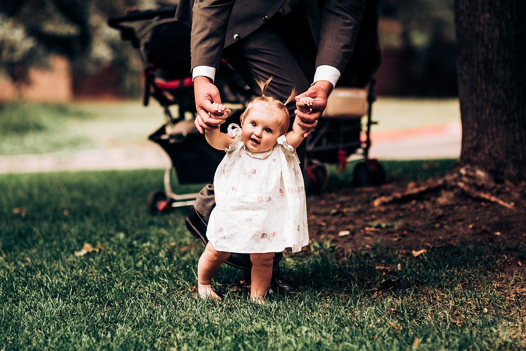 Small child in white dress taking first steps while holding adult hands.
