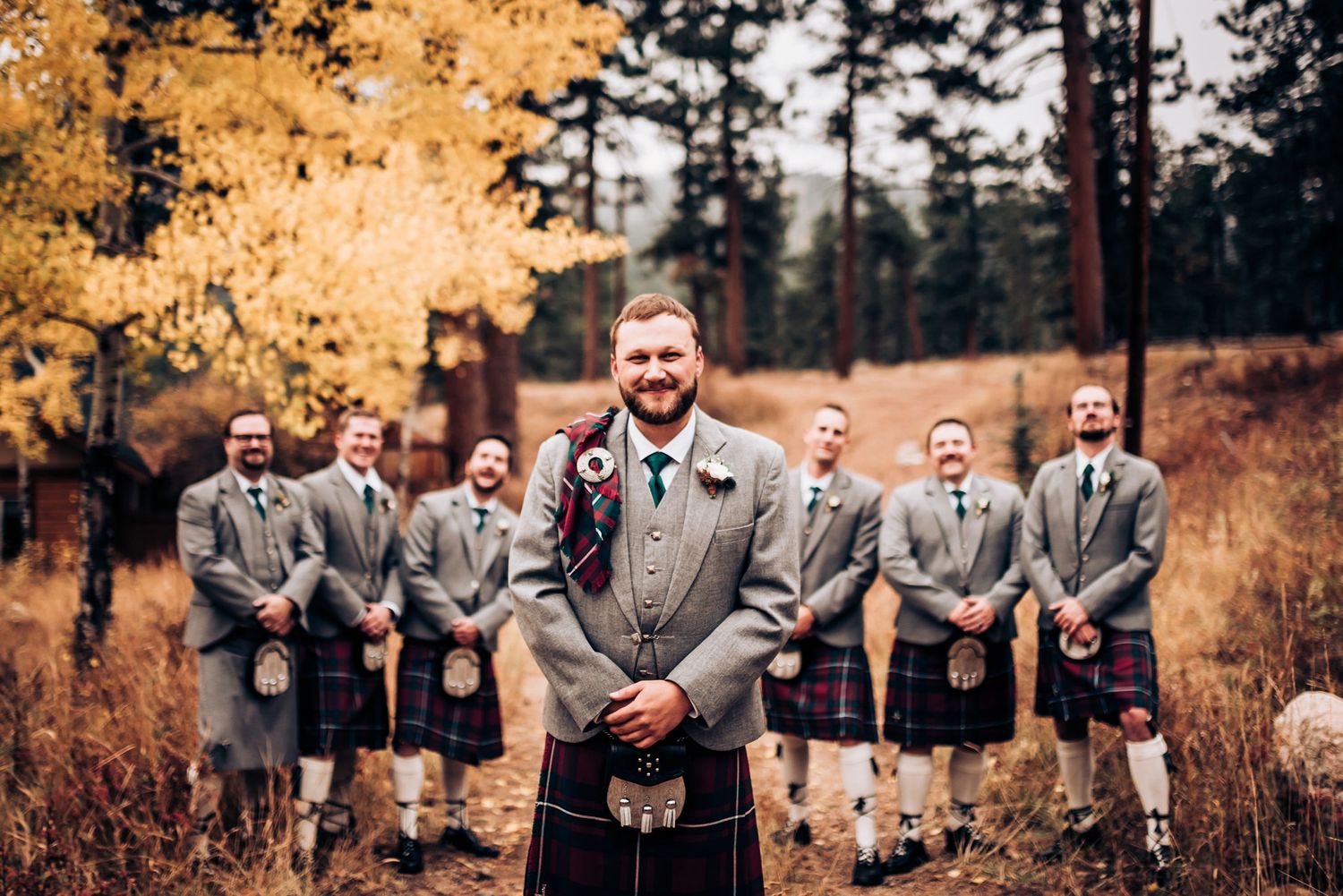 Groomsmen in grey kilts stand behind groom for formal wedding photo in autumn setting.