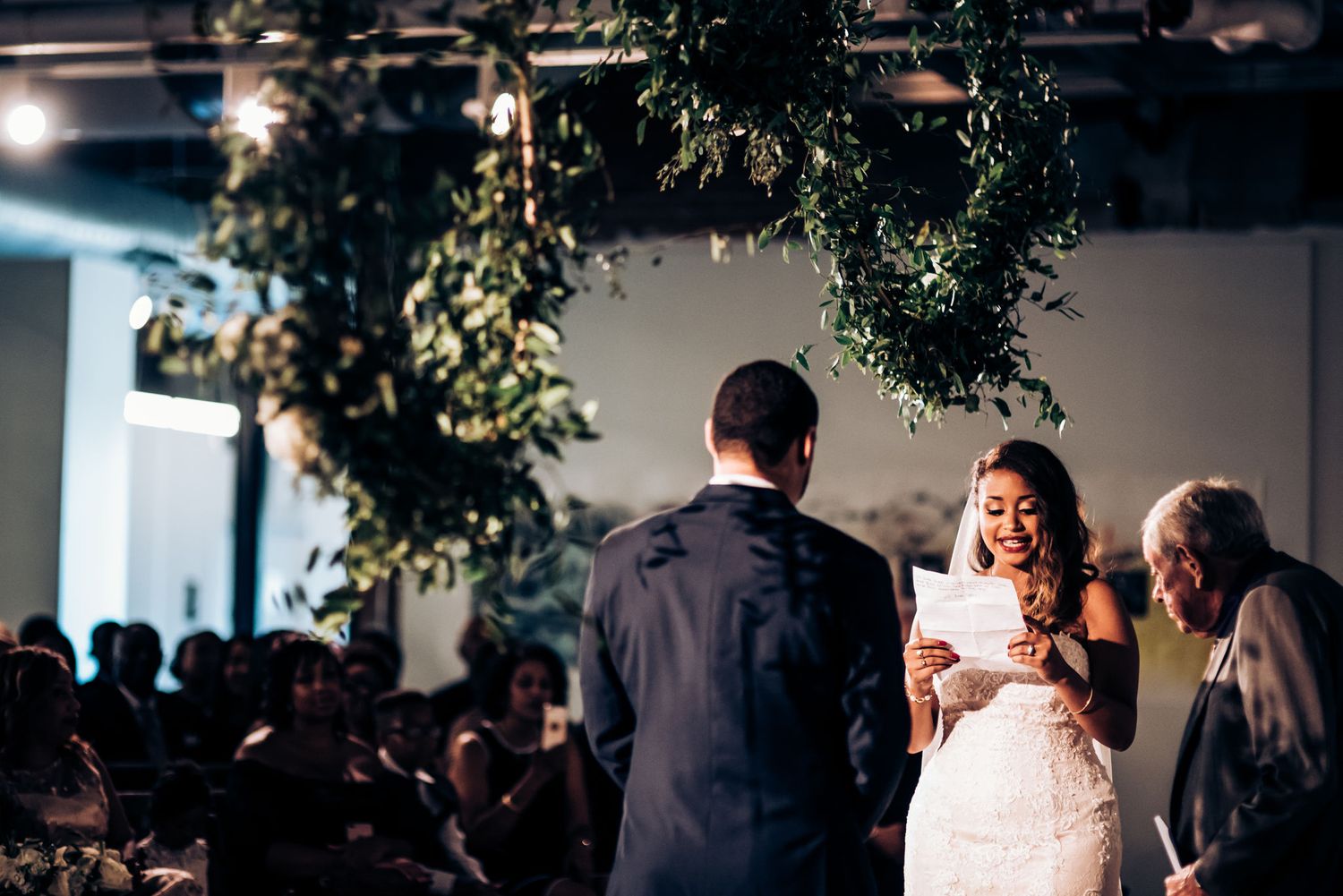 A bride reads her vows during an indoor ceremony decorated with hanging greenery.