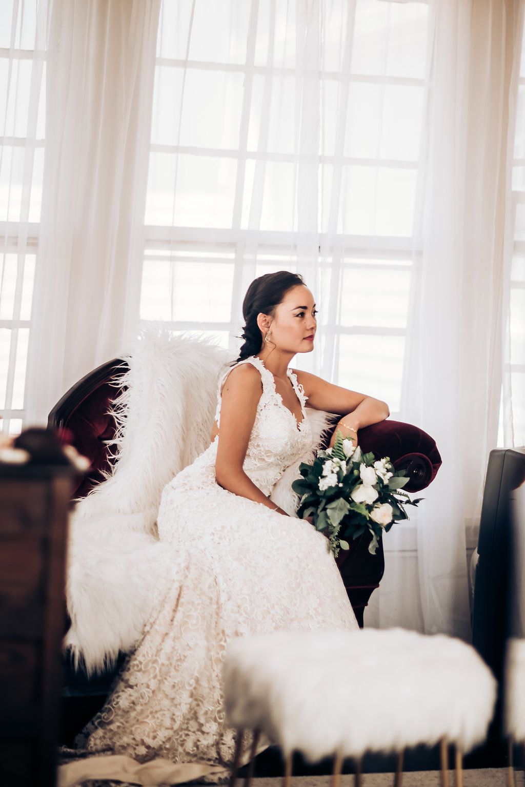 Bride in lace gown sits elegantly on dark velvet couch holding white floral bouquet.