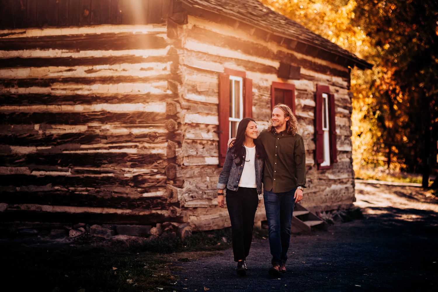 Couple walks together along path beside rustic log cabin during golden hour.