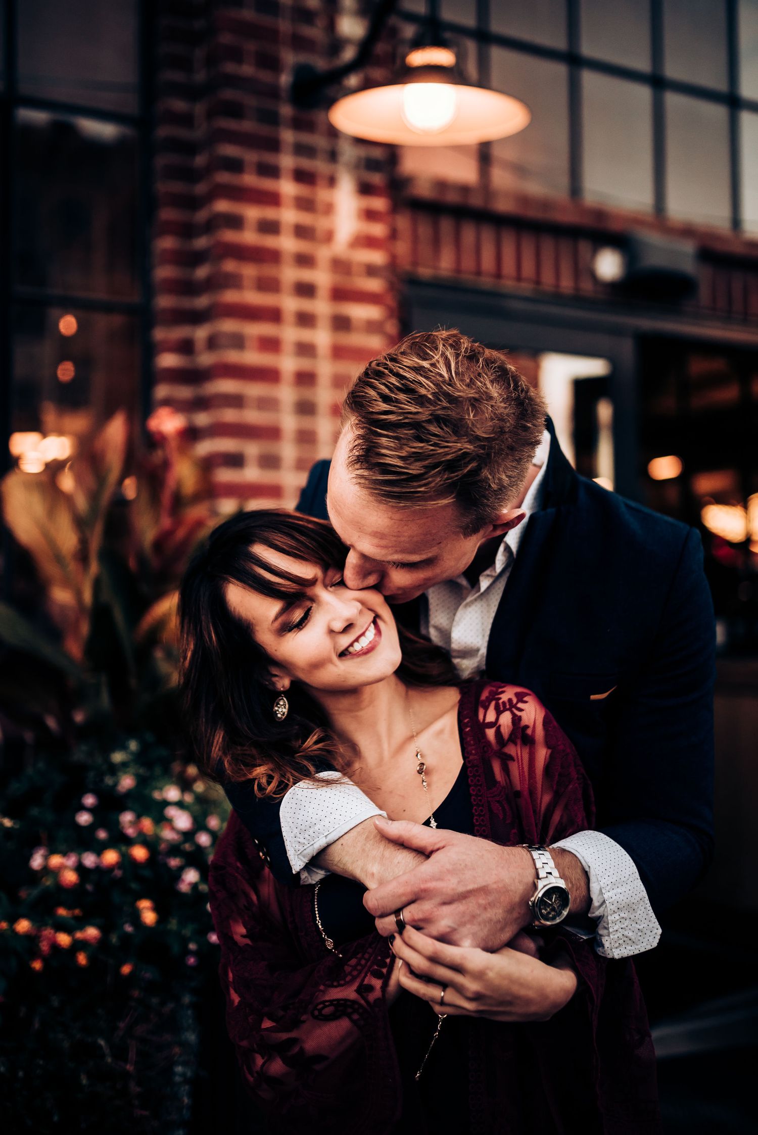 A romantic couple shares an intimate embrace under warm lighting against a brick wall backdrop.