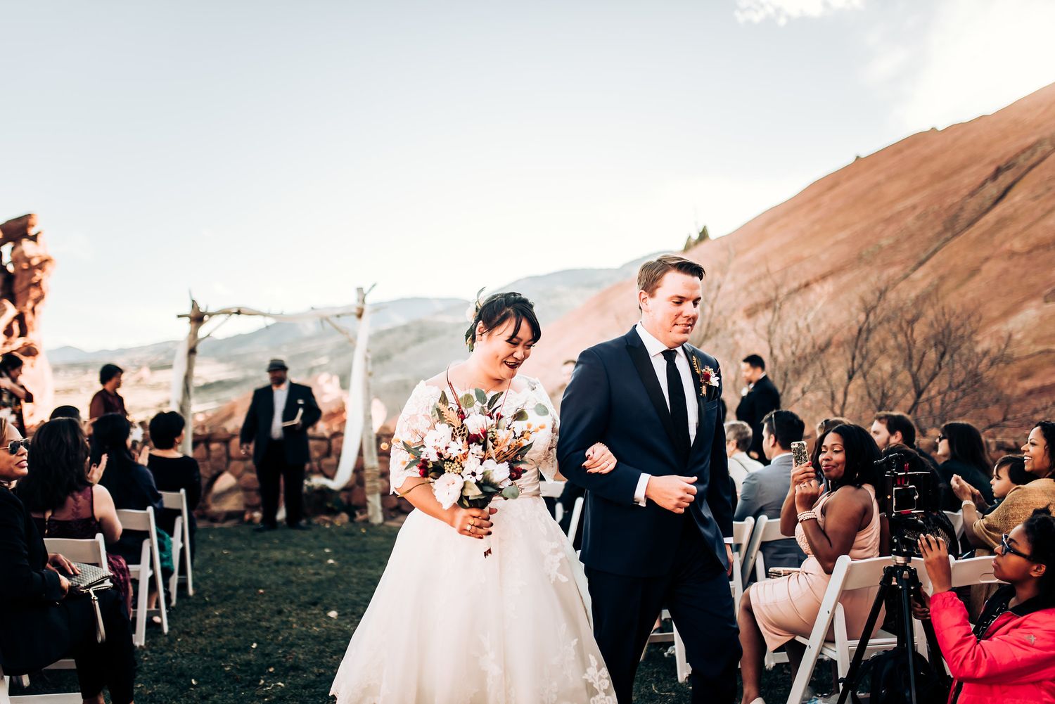Couple walks down aisle at outdoor mountain wedding with guests seated on white chairs.