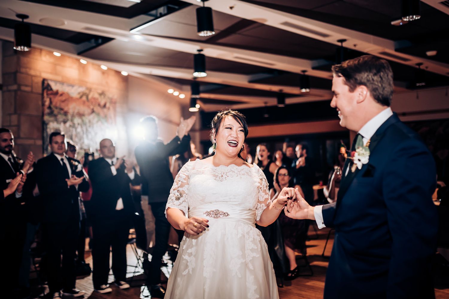 A wedding couple shares a lively dance moment in a warmly lit reception hall with guests looking on.