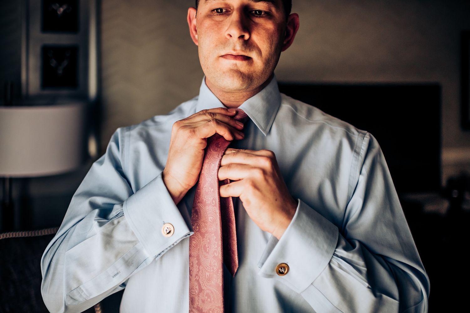 A close-up shot of hands adjusting a salmon-colored tie with a light blue dress shirt.