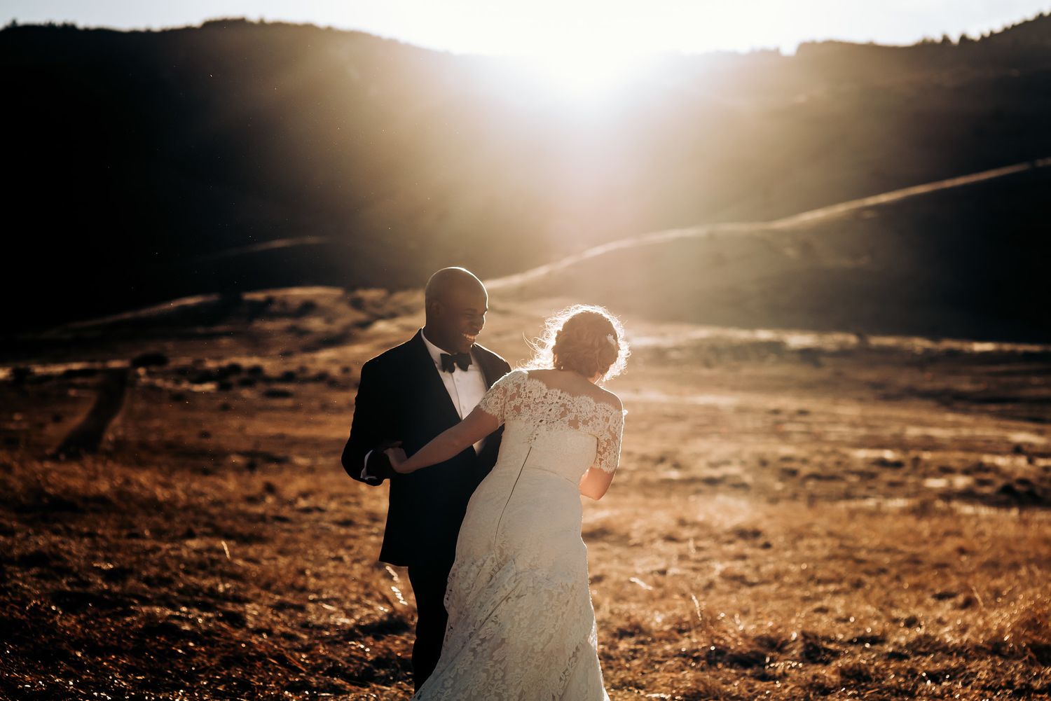 Couple walks hand in hand through golden mountain meadow at sunset.