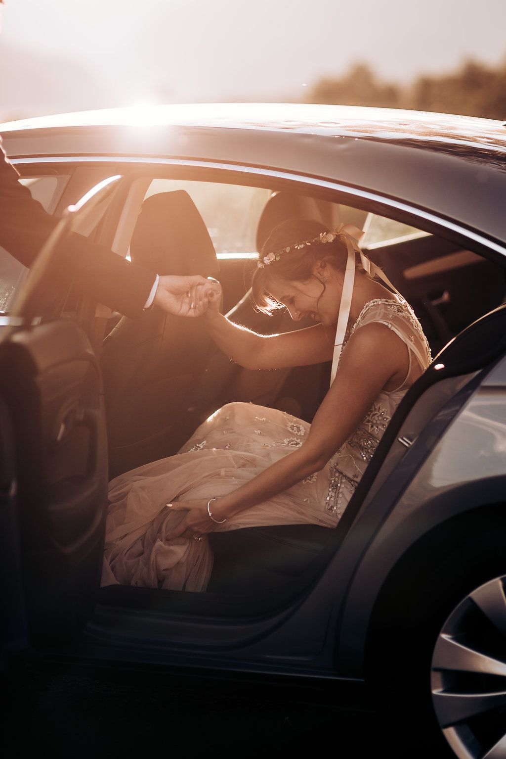 A bride in a beaded gown getting into a car during sunset with light flare through the window.
