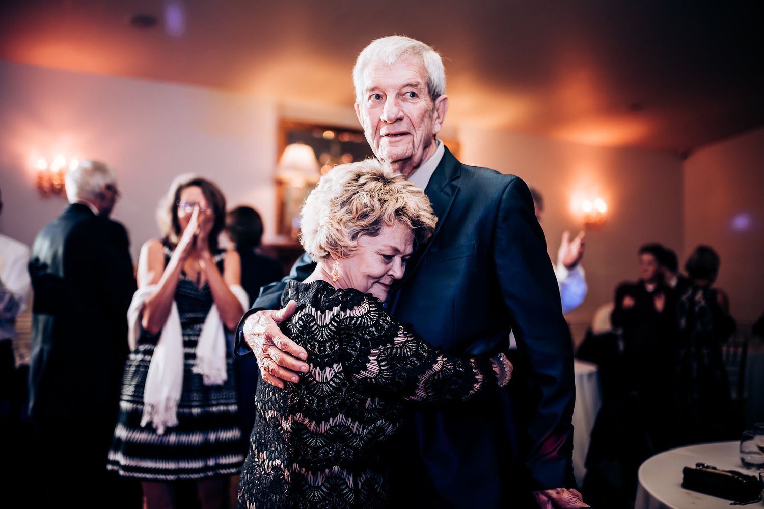 Dancers embrace in a heartfelt moment on a formal event dance floor with warm lighting and guests in the background.