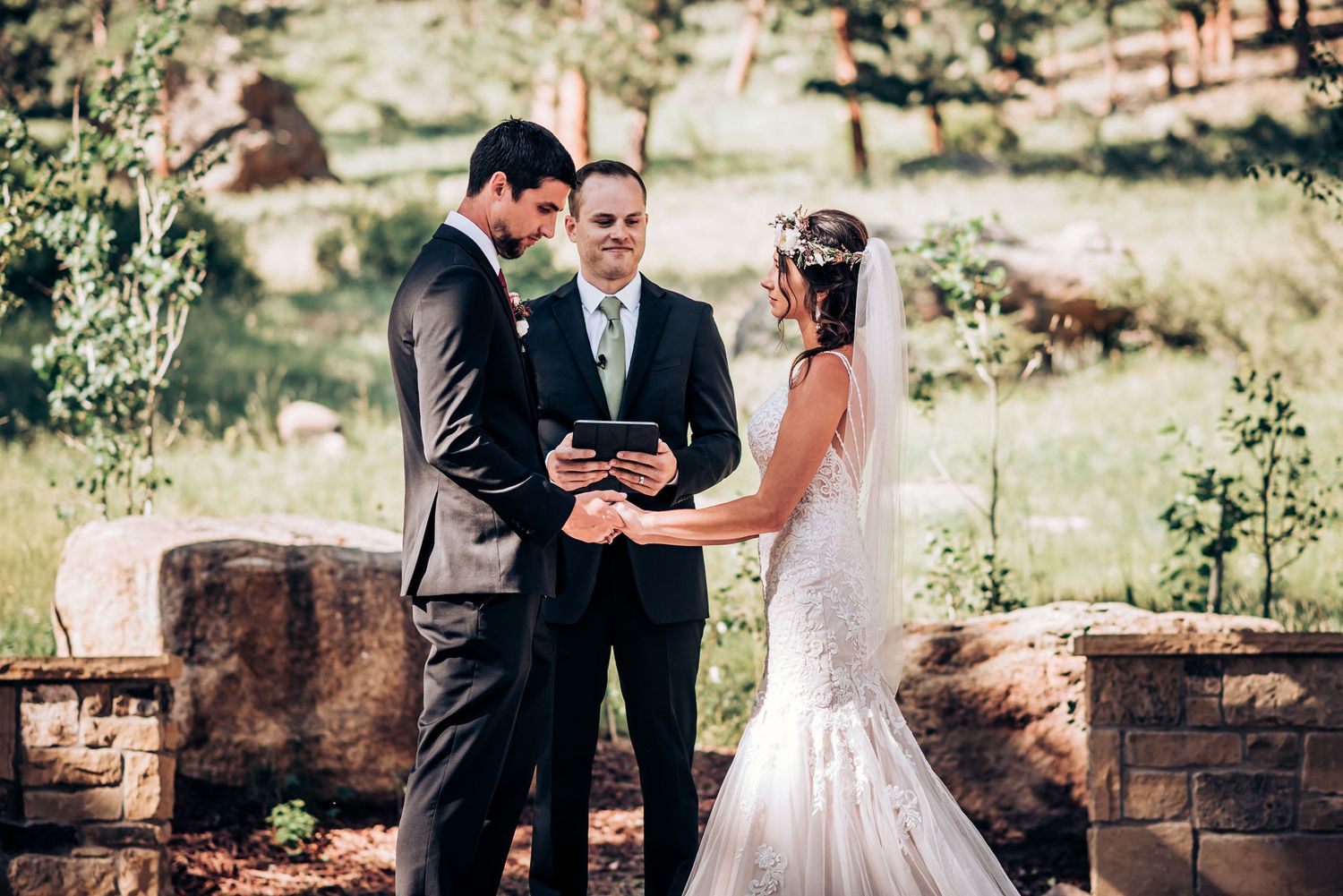 A wedding ceremony takes place under a rustic wooden pergola in a mountain forest setting.