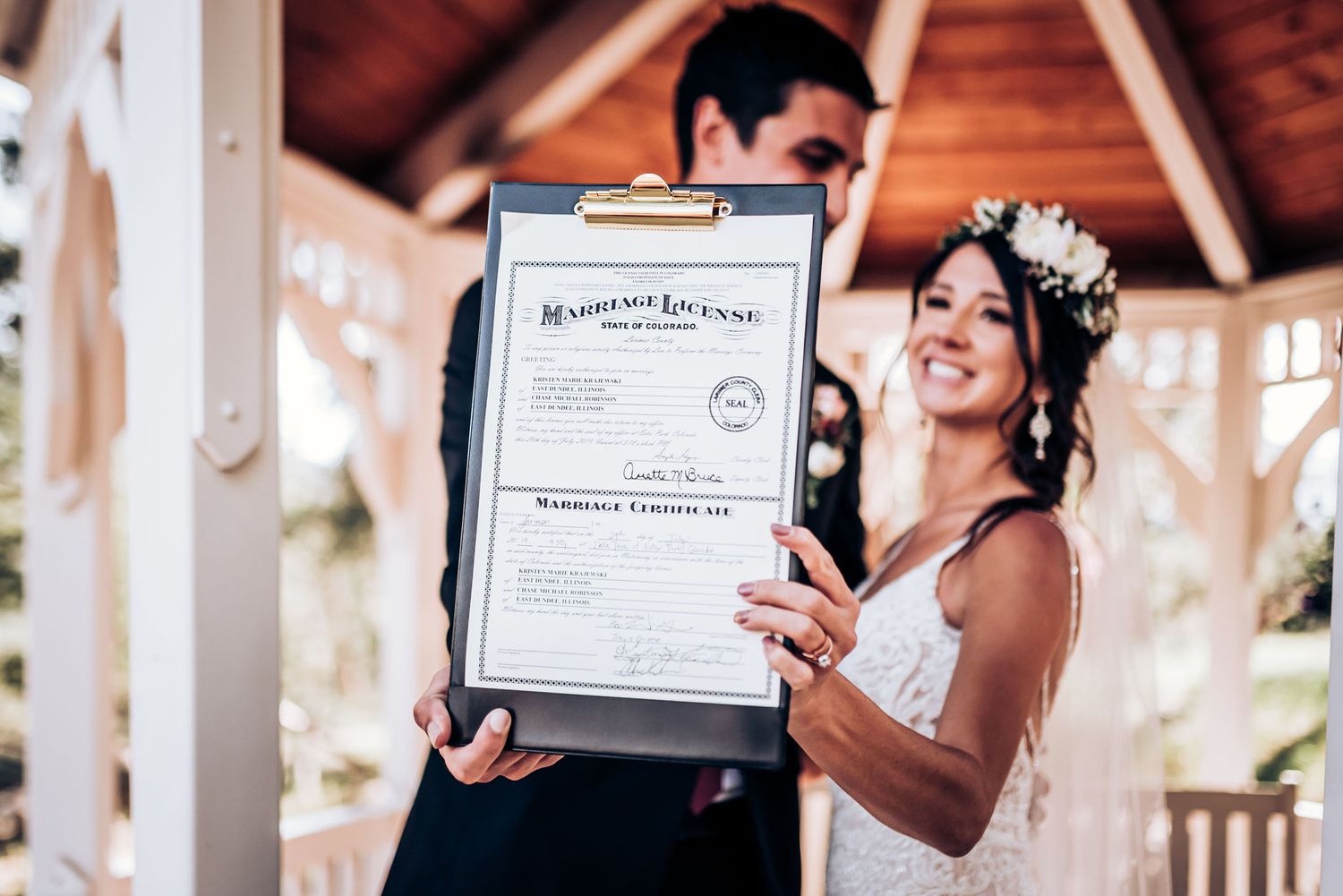 A couple joyfully showing their marriage certificate in an outdoor pavilion.